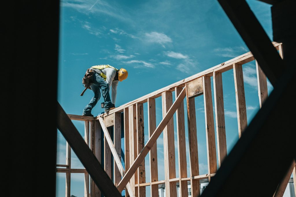 Construction worker wearing hard hat, balancing on wooden beam, high up on a building, against a blue sky.