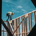 Construction worker wearing hard hat, balancing on wooden beam, high up on a building, against a blue sky.